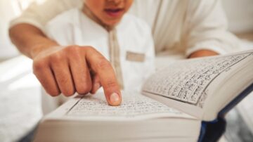 A close-up of a child's hand pointing at text in a book, with an adult's hand partially visible in the background. The child appears focused on reading, showcasing a moment of learning. The book features intricate calligraphy, suggesting it may be a religious text.