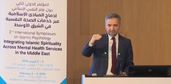 رجل يتحدث في مؤتمر دولي عن إدماج الممارسات الإسلامية في خدمات الصحة النفسية، مع وجود لافتة للمؤتمر توضح التفاصيل. التاريخ: 4 فبراير 2026. المكان: قاعة المحاضرات.