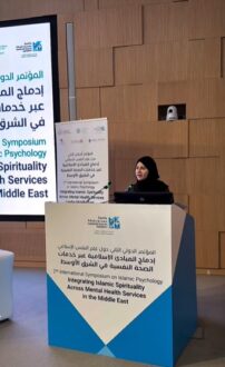 امرأة ترتدي حجابًا تتحدث في مؤتمر حول تكامل الروحانية الإسلامية مع خدمات الصحة النفسية في الشرق الأوسط، مع وجود لافتة توضح تفاصيل المؤتمر في الخلفية.