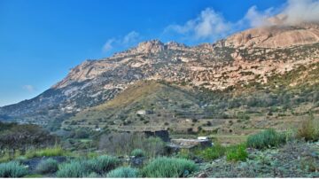 Scenic landscape of rugged mountains under a blue sky, featuring rocky cliffs and lush greenery in the foreground. The terrain includes terraced fields and distant structures, showcasing natural beauty and mountainous terrain.