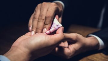 تبادل نقود بين شخصين، حيث يقوم أحدهما بإعطاء ورقة نقدية للآخر، في بيئة ذات إضاءة منخفضة. يُظهر المشهد التركيز على اليدين والنقود، مما يعكس عملية مالية أو اتفاق مالي.