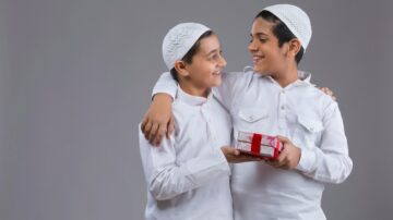 شقيقان يرتديان قمصان بيضاء وطاقيات، يبتسمان ويتعانقان، أحدهما يحمل هدية مزينة بشرائط حمراء. الخلفية رمادية. تعكس الصورة فرحة المشاركة والاحتفال.