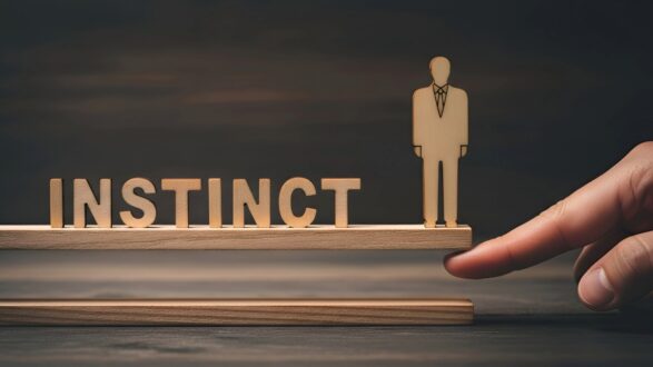 صورة توضح كلمة "INSTINCT" مكتوبة بأحرف خشبية، مع وجود تمثال خشبي لرجل يرتدي بدلة، بينما يلمس إصبع يد إنسان قاعدة الكلمة. تعكس الصورة مفهوم الحدس في اتخاذ القرارات.