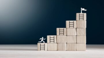 صورة توضح كتل خشبية مرتبة على شكل درجات، مع رمز لشخص يتسلق إحدى السلالم. في أعلى الدرجات، توجد علم صغير، مما يرمز إلى تحقيق الأهداف والطموحات.