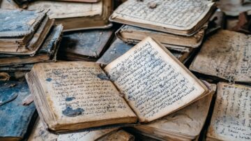 كتب قديمة مكدسة فوق بعضها البعض، مع صفحات مفتوحة تظهر نصوصًا عربية مكتوبة بخط يدوي، تعكس تاريخًا وثقافة غنية، تغطيها طبقة من الغبار.