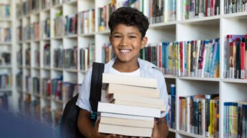 طفل مبتسم يحمل مجموعة من الكتب في مكتبة مليئة بالرفوف المليئة بالكتب.