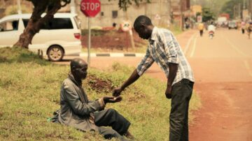 رجل شاب يقدم المساعدة لرجل مسن يجلس على جانب الطريق، مع وجود علامة توقف في الخلفية. تعكس الصورة مشهدًا إنسانيًا يظهر التعاطف والعطاء في المجتمع.