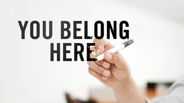 يد تحتوي على قلم تكتب عبارة "أنت تنتمي هنا" بخط عريض، مع خلفية بسيطة تعزز الرسالة الإيجابية.