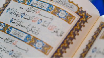 Close-up view of an open Quran displaying Arabic text with intricate gold and blue decorative borders. The page features verses from the Quran, highlighting the beauty of Islamic calligraphy.