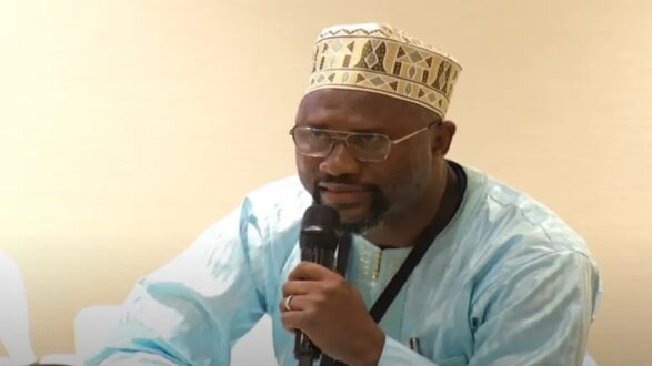 رجل يرتدي زي تقليدي أزرق وقبعة مزخرفة، يتحدث إلى ميكروفون خلال حدث رسمي. تظهر خلفيته جدران بيضاء، مما يبرز تركيزه على التحدث.