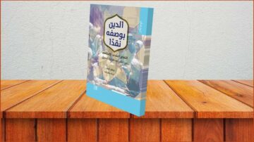 كتاب بعنوان "الحين بوصفه نقداً" يتناول الفكر النقدي المعاصر، موضوعه يظهر على غلافه الملون. الكتاب موضوع على سطح خشبي، مما يضيف لمسة دافئة تعكس البيئة الثقافية.