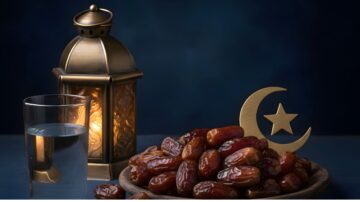 A plate of dates sits beside a glass of water and a decorative lantern, with a gold crescent moon and star symbol in the foreground. The background features a deep blue hue, creating a serene ambiance. Ideal for Ramadan themes and Iftar celebrations.