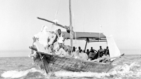 قارب خشبي تقليدي يبحر في البحر، مع مجموعة من الرجال على متنه. يظهر بعضهم وهم يعملون بينما يجلس الآخرون. المياه هادئة، والسماء واضحة. صورة تاريخية تعكس الحياة البحرية.