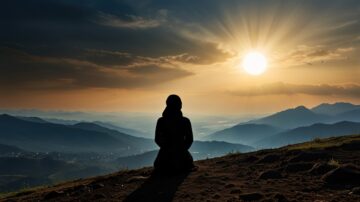 Silhouette of a person sitting on a hillside at sunset, with mountains and valleys in the background. The sun is low on the horizon, casting a warm glow across the landscape.