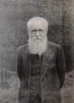 رجل مسن ذو لحية وشعر أبيض، يرتدي نظارات وقميصًا أسود مع سترة، يقف في غابة. تعكس الصورة طابعًا تاريخيًا وجوًا هادئًا.