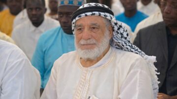 رجل مسن يرتدي عقالاً وعباءة بيضاء، يجلس بين مجموعة من الرجال في مناسبة اجتماعية، مع تعبير هادئ على وجهه، بينما يرتدي الآخرون ملابس ملونة متنوعة.