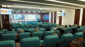 قاعة مؤتمرات حديثة تحتوي على مقاعد زرقاء، حيث يجلس جمهور متنوع يستمع إلى محاضرة تُعرض على شاشة كبيرة. المتحدث يظهر في الخلفية على الشاشة، مع وجود كاميرات تسجل الحدث.