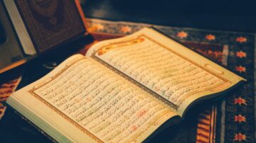 كتاب مفتوح مع صفحات تحتوي على نصوص عربية، يظهر الآيات القرآنية بتنسيق جميل. خلفية الصورة سجادة تقليدية، مما يضيف جوًا روحانيًا.