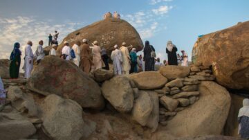 Crowd of people in traditional attire gathered around large boulders at a historic site. Entrance of the Cave of Thawr on Jabal Thawr in Makkah