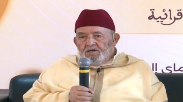 رجل مسن يرتدي جلبابًا تقليديًا وقبعة حمراء، يتحدث إلى ميكروفون خلال حدث. الخلفية تحتوي على نص عربي يشير إلى موضوع النقاش.