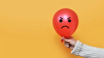 Red balloon with an angry face, held by a hand against a bright yellow background. The balloon features a frowning expression, conveying emotions of frustration or anger.