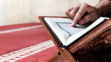 A person’s hand points to a page of an open Quran on a wooden stand, set against a red patterned carpet background.