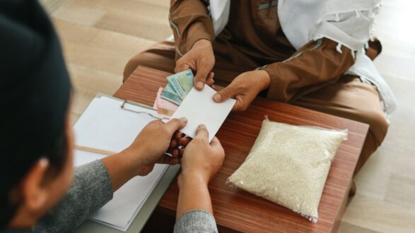 شخصان يجلسان على طاولة خشبية، أحدهما يسلم ظرفاً مالياً للآخر، بينما توجد كيس من الأرز على الطاولة. يظهر في الخلفية أوراق وأموال. تعكس الصورة عملية تبادل أو مساعدة مجتمعية.