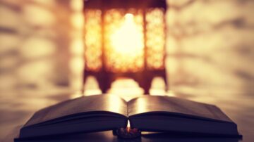 كتاب مفتوح أمام فانوس مضيء، يخلق جوًا دافئًا وهادئًا. يركز الضوء على صفحات الكتاب، مما يبرز تفاصيل النص. الخلفية ضبابية مع تأثيرات ضوئية ناعمة تضيف لمسة من السحر.