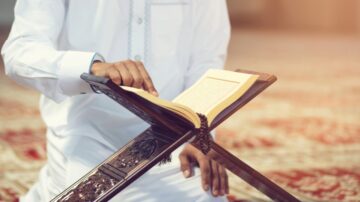 رجل يرتدي ثوبًا أبيض يجلس على سجادة في مكان الصلاة، وهو يقرأ القرآن الكريم من مصحف موضوع على حامل كتب خشبي.