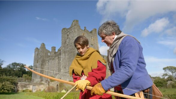 شاب ومرشد يمران بتدريب على رماية السهام أمام قلعة قديمة. السماء زرقاء مع بعض الغيوم، والقلعة تظهر في الخلفية. الشاب يرتدي ملابس تقليدية ملونة بينما يركز على توجيه السهم.