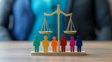 ميزان يتوازن عليه أشخاص ملونون، يمثل العدالة والمساواة. الخلفية ضبابية مع شخص يرتدي بدلة، مما يعكس بيئة مهنية.