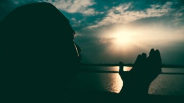 رجل يرفع يديه للدعاء أمام شمس غاربة تعكس أشعتها على مياه النهر الهادئة، مع سماء ملبدة بالغيوم.