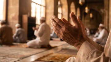 يدٌ مرفوعةٌ بالدعاء في مسجد تقليدي، مع مجموعة من المصلين في الخلفية. تُظهر الصورة أجواء السكينة والتركيز أثناء الصلاة.