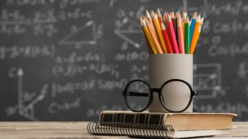 قلم ملون في وعاء على طاولة مع نظارات وقاموس مفتوح، خلفية تحتوي على معادلات رياضية مكتوبة على السبورة.