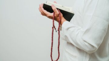 شخص يحمل كتابًا دينيًا في يد وسبحة في اليد الأخرى، يرتدي ملابس بيضاء، مع خلفية بسيطة.