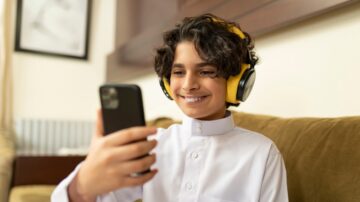 طفل يرتدي سماعات رأس صفراء ويبتسم أثناء استخدام هاتفه الذكي في غرفة معيشة مريحة.