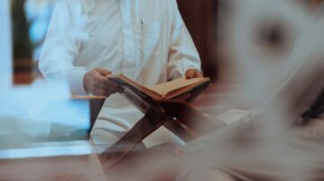 رجل يرتدي ثوبًا تقليديًا يحمل مصحفًا مفتوحًا، يجلس أمام منصة قراءة. يظهر خلف زجاج شفاف، مما يضيف لمسة فنية للصورة.