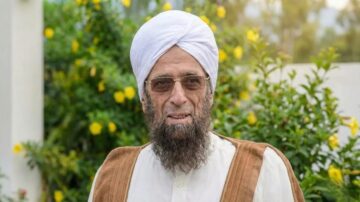 رجل مسن يرتدي عمامة بيضاء ونظارات، يظهر بابتسامة في بيئة طبيعية مزهرة مع أزهار صفراء وخلفية خضراء.