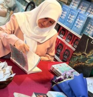 امرأة ترتدي الحجاب تكتب توقيعها على كتاب في حدث توقيع كتب، مع وجود كتب ملونة مرتبة خلفها وزهور على الطاولة.