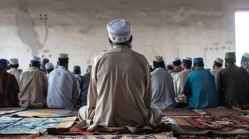 A group of men sitting in a prayer space, facing a wall, with a focus on one man in traditional attire and a white cap. The setting suggests a communal prayer or religious gathering.