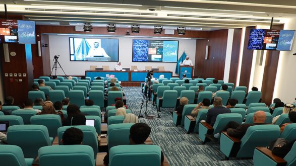 اجتماع رسمي في قاعة مؤتمرات حديثة مع جمهور كبير يجلس على مقاعد زرقاء. يتواجد متحدث على المنصة يعرض محتوى عبر شاشة كبيرة. تم إعداد كاميرات لتغطية الحدث.