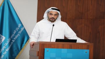 رجل يرتدي الزي التقليدي الإماراتي يقف خلف منصة يتحدث في حدث رسمي، مع وجود علم مركز الأبحاث والدراسات السياسية في الخلفية.