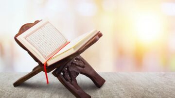 كتاب مفتوح على حامل خشبي، يظهر في خلفية مضيئة، مما يضفي جوًا هادئًا ومناسبًا للتأمل والقراءة.