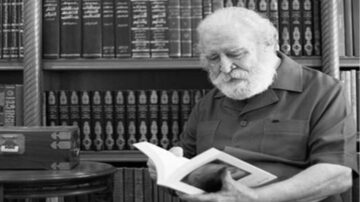 رجل مسن ذو لحية وشعر أبيض يجلس في مكتبة تحتوي على رفوف مليئة بالكتب. يقرأ كتابًا بتركيز، بينما يظهر في الخلفية مجموعة من الكتب العربية.