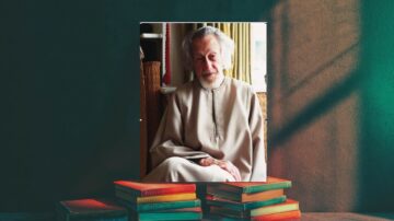 رجل مسن ذو لحية رمادية يجلس مبتسمًا في غرفة مضاءة بشكل دافئ، يرتدي جلبابًا بسيطًا. خلفه نافذة تتيح دخول الضوء الطبيعي، وهناك مجموعة من الكتب الملونة موضوعة أسفل الصورة.