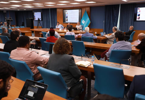 اجتماع يناقش مواضيع متنوعة في قاعة مؤتمرات، مع وجود متحدثين على المنصة ومواضيع معروضة على الشاشة. الحضور يتوزع بين رجال ونساء، مع وجود أجهزة كمبيوتر محمولة وأجهزة تسجيل.