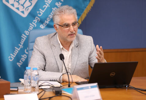 رجل يتحدث في اجتماع، يجلس أمام جهاز كمبيوتر محمول، مع زجاجتين من الماء على الطاولة. خلفه لافتة تحمل شعار مركز الأبحاث العربي.