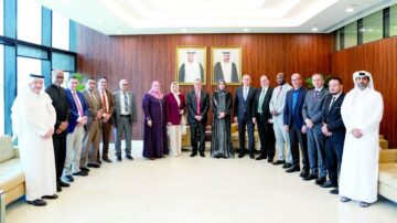 اجتماع رسمي لمجموعة متنوعة من الأفراد في غرفة اجتماعات حديثة، مع صور لرجلين في الإطار خلفهم. الحضور يتضمن رجال ونساء يرتدون ملابس رسمية، ويبدو عليهم التفاعل الإيجابي.