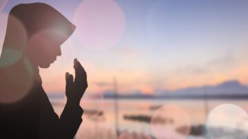 Silhouetted person in modest Islamic clothing, with hands raised in prayer, at sunrise against a lake, representing Haya, spiritual purity, and Divine proximity