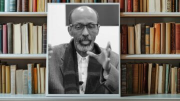 رجل يتحدث بحماس مع تعبيرات وجه واضحة، يرتدي نظارات ووشاح، في إطار محادثة أو مناقشة. خلفه مكتبة مليئة بالكتب، تعكس أجواء أكاديمية وثقافية.